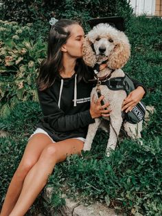 a woman is kissing her poodle while sitting on the ground in front of some bushes