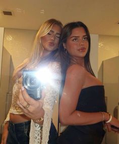 two beautiful young women standing next to each other in front of a mirror holding a camera