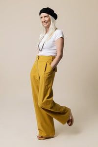 a woman in white shirt and yellow pants standing with her hands behind her back, smiling at the camera