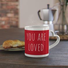 a coffee mug with the words you are loved on it next to a croissant