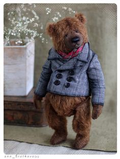 a teddy bear dressed in a coat and scarf standing next to a vase with flowers