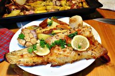 a white plate topped with fish next to a casserole filled with potatoes and veggies