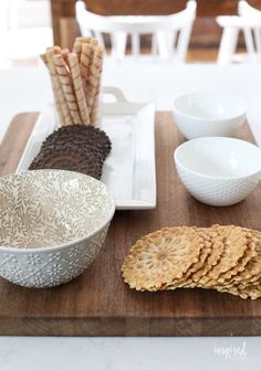 a wooden cutting board topped with white bowls and plates filled with crackers on top of it