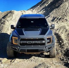 the front end of a gray truck driving down a dirt road next to a mountain