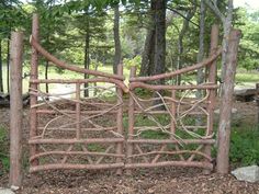 a wooden gate made out of branches in the middle of some grass and dirt with trees behind it