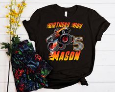 a black shirt with an orange and yellow monster truck on it's chest, next to some flowers