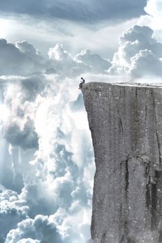 a man standing on the edge of a cliff with clouds in the sky behind him