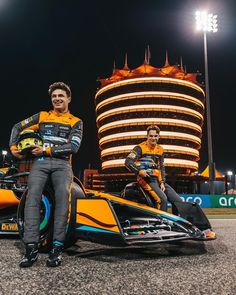 two men sitting on top of a race car in front of a tall building at night