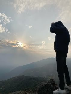 a man standing on top of a mountain looking at the sun in the distance with his hands behind his head