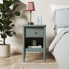 a nightstand with a lamp on top of it next to a bed and potted plant