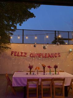 a table set up for a party with lights strung above it and flowers in vases on the table