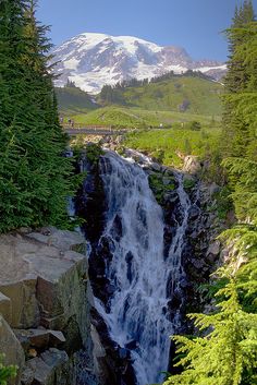 a large waterfall surrounded by trees and snow capped mountain in the background with blue sky