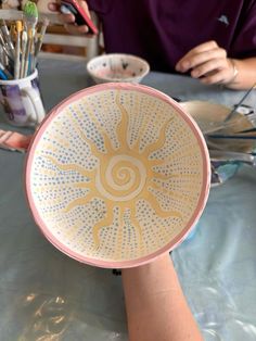 a person sitting at a table with a bowl on it's head and paintbrushes in the other hand