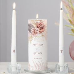 two white candles with pink flowers on them next to some glass vases and a candle holder