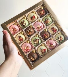 a hand holding a box filled with assorted decorated christmas baubles on top of a white tile floor