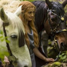 a woman standing next to two horses in the woods with long red hair on her head