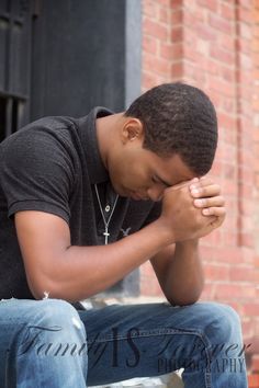 a young man sitting on a bench in front of a brick building holding his hands to his face