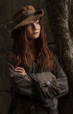 a woman with long red hair wearing a brown hat and jacket standing in front of a tree