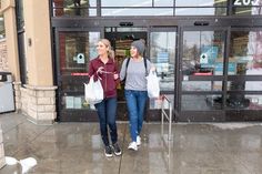 two women walking down the sidewalk in front of a building with snow falling on them