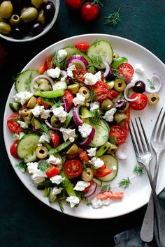 a white plate topped with cucumbers, tomatoes and olives next to silverware