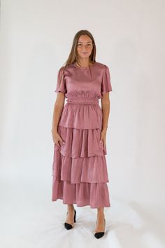 Discover the charm of the Marisa Tiered Dress, a stunning dusty pink midi dress perfect for any occasion. This dress features a beautifully tiered design that adds depth and graceful movement. Crafted from high-quality, soft satin-finished fabric, the Marisa Dress ensures a comfortable fit throughout the day. The round neckline, flattering smocked waistline, and flutter sleeves provide a classic touch, while the tiered layers create a playful and sophisticated look. The delicate mauve color adds Rose Satin Dress, Graceful Movement, Holiday Shoes, Tiered Midi Dress, Mauve Color, Pink Midi Dress, Tiered Dress, Flutter Sleeves, Skirt Top