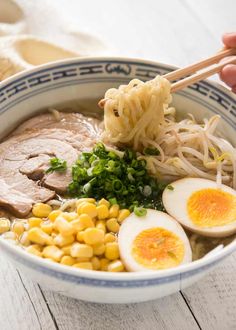 someone holding chopsticks over a bowl of ramen with boiled eggs and corn