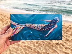 a hand holding up a painting of a mermaid on the beach with waves in the background