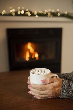 an older woman holding a cup of hot chocolate in front of a fireplace