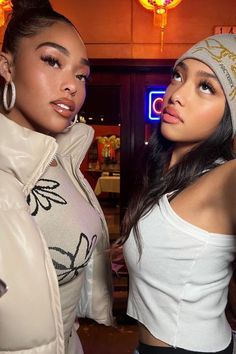 two beautiful women standing next to each other in front of a neon lit bar and looking at the camera