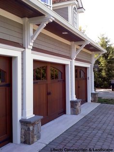 two brown garage doors sitting on the side of a white and gray house with brick walkway