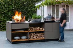 a man standing in front of a grill with firewood on the side and bowls next to it