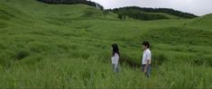 two people standing in tall grass looking at the sky and hills behind them on a cloudy day