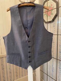 This classic vest looks grey from a distance, but up close it has black, white and medium blue fibres in the polyester blend fabric. It has the original buttons and two front pockets. The back and lining are made from silky bluish grey fabric. The measurements, taken with the vest lying flat, are: shoulder to shoulder, 13 inches; armpit to armpit, 22 inches; length, 25 inches in front and 21 inches in back; bottom edge, 22 inches. In very good condition. Affordable Gray Fitted Vest, Classic Tailored Gray Vest, Classic Gray Vest For Work, Fitted Gray Vest With Pockets, Gray Tailored Vest For Business, Gray Sleeveless Vest Outerwear, Fitted Gray Vest For Business, Gray Fitted Vest For Business, Classic Gray Formal Vest