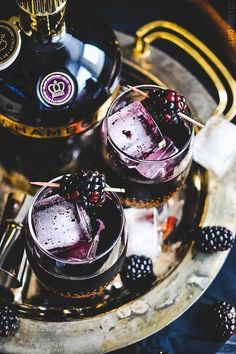 two glasses filled with ice and blackberries on top of a metal tray next to a bottle