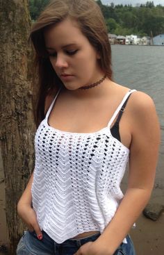 a young woman standing next to a tree near the water with her hands in her pockets