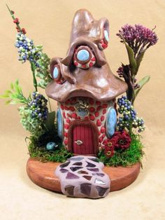 a small house made out of clay sitting on top of a wooden base with flowers and plants around it