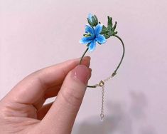 a hand holding a tiny blue flower with green leaves and chain attached to it's side