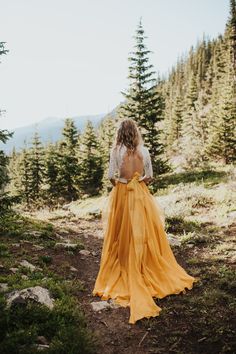 a woman in a long yellow dress walking through the woods with her back to the camera