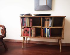 a wooden book shelf with books on it in front of a chair and wall hanging