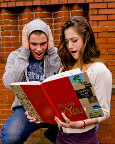 a man and woman sitting on the ground reading a book with their hands behind them