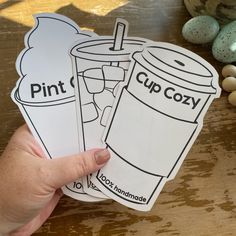 hand holding up two cup cozy stickers on top of a table next to eggs