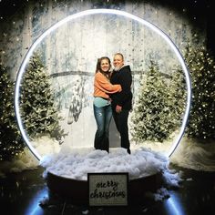 two people standing in front of a snow globe with christmas trees and lights behind them