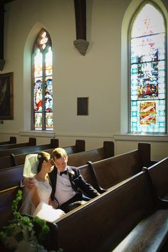 a bride and groom sitting in the pews of a church, with stained glass windows behind them