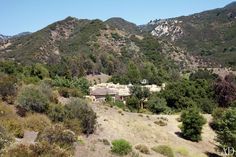 a house in the middle of some trees and bushes on a hill with mountains in the background