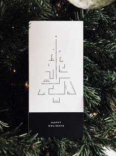 a christmas tree ornament hanging from the top of a fir tree with a black and white card that says happy holidays