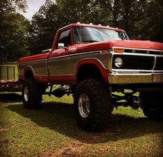 an old red truck parked on top of a grass covered field with trees in the background