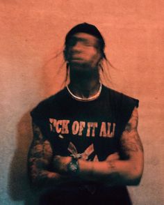 a man with long hair and tattoos standing in front of a wall holding his arms crossed