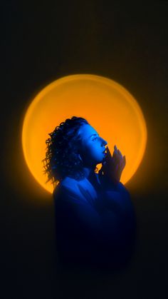 a woman with curly hair standing in front of a yellow light and holding her hand to her face