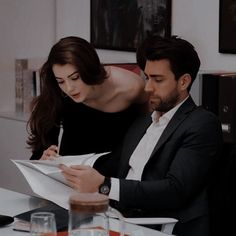 a man and woman sitting at a table with papers in front of them, looking at each other