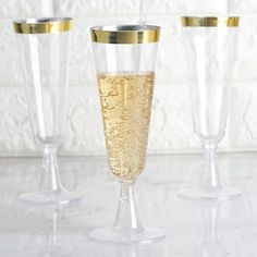 three champagne flutes with gold rims are lined up on a white countertop in front of a brick wall